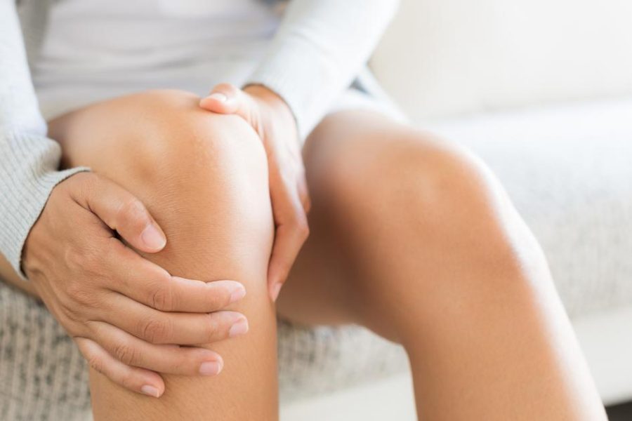 Close-up of a person sitting on a couch, holding their knee with both hands, possibly indicating pain or discomfort.